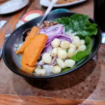 Amar Peruvian Kitchen - Ceviche De Pescado (fish.) Outstanding - A black bowl contains ceviche with corn, sliced red onions, fresh lettuce, and slices of sweet potato, placed on a wooden table. In the background, there are additional plates and a person partially visible, indicating a dining setting at a restaurant. - Peruvian Near Me - Forest Hills, New York