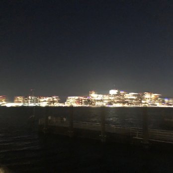 Hyatt Regency Boston Harbor - You would night from the Hyatt Regency Harbor - A nighttime scene showing a city skyline with brightly lit buildings across a body of water. The image is blurry, making the buildings appear as streaks of light. A pier or railing is visible in the foreground, indicating the photo was taken from a waterfront location. - Hotels Near Me - Boston, Massachusetts