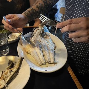 Andros Taverna - A person with tattooed arms is using a fork and knife to cut a cooked fish on a white plate at a restaurant. The table has additional dishes, including a plate with fish bones, a small bowl of oil, and condiments. Other diners and restaurant interior elements are visible in the background. - Greek Near Me - Chicago, Illinois