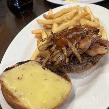 George's - A white oval plate holds a serving of golden French fries, a cheeseburger topped with crispy bacon and caramelized onions, and a toasted bun with melted cheese. In the background, a glass of dark soda and additional breakfast items, including pancakes, bacon, scrambled eggs, and syrup, are visible on a dark wooden table. - Diners Near Me - Brooklyn, New York