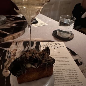 The Guest - The image shows a dessert with a rectangular base topped with dark chocolate and moss-like garnish, placed on a small white plate. A tall, elegant glass of white wine and a glass of water are positioned behind the dessert on a restaurant table. The table has a welcome note and a menu, with a background of paneled walls, indicating an indoor dining setting. - Modern European Near Me - Denver, Colorado