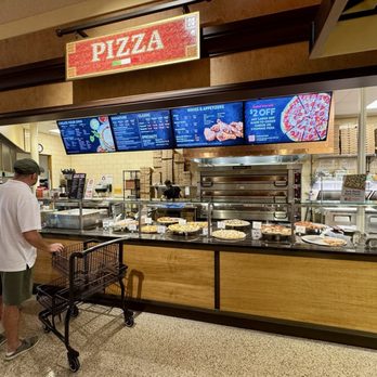 Wegmans - Pizza Bar - A pizza counter inside a restaurant features digital menu boards displaying various pizza options and prices. The counter has a glass display with several pizzas on trays, and pizza boxes are stacked behind it. Customers are seen waiting in line, with one person pushing a shopping cart. The background shows part of the restaurant interior with a tiled wall and ceiling lights. - Grocery Near Me - Norwalk, Connecticut