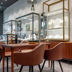 Adam Goldschmiede - Braune Ledersessel um Holztisch vor beleuchteten Schmuckvitrinen. Elegante Pendelleuchten. Grauer Teppich. Hochwertige Präsentation