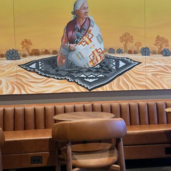 Starbucks - beautiful artwork - A mural depicts an elderly woman sitting on a black and white patterned rug in a desert scene with trees and a yellow sky. - Coffee & Tea Near Me - Gallup, New Mexico