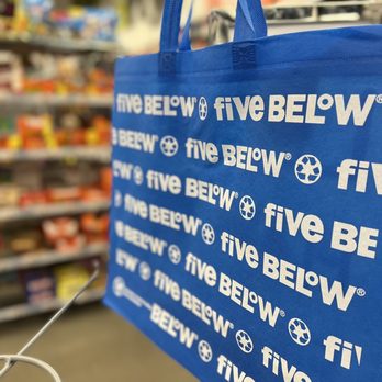 Five Below - A blue reusable shopping bag featuring the Five Below logo and branding in white is prominently displayed in the foreground. The bag is held up in a retail store aisle, with shelves stocked with various products visible in the background. The image captures the store environment, emphasizing the store's branding and product display area. - Discount Store Near Me - East Brunswick, New Jersey