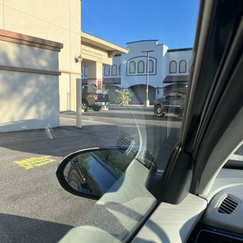 CVS Pharmacy - Busy Drive-Thru - A parking lot scene featuring a CVS Pharmacy with a drive-thru, viewed from inside a vehicle. The photo captures the building's exterior, including the drive-thru pharmacy sign, parked cars, and a clear blue sky. The vehicle's side mirror and part of the dashboard are visible in the foreground, indicating the photo was taken from the driver's seat. Shadows from nearby trees are cast on the building's wall, and the overall setting appears calm and sunny. - Drugstores Near Me - Chandler, Arizona