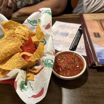 El Bruno's - $5 chips - A basket filled with yellow tortilla chips, including a red chip, is placed on a restaurant table lined with branded paper. Next to the basket is a small bowl of red salsa with visible green herbs. The table also has a menu and a glass of water, with partially visible people seated nearby. The setting suggests a casual dining environment, likely Mexican or Tex-Mex cuisine. - Mexican Near Me - Albuquerque, New Mexico