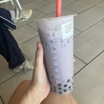 Tea Bliss - Taro Boba! - A person is seated indoors, holding a large plastic cup of bubble tea with a pink straw. The drink contains tapioca pearls at the bottom and appears to be a taro milk tea flavor. The background shows tiled flooring and part of a white chair, with a person wearing sneakers and dark shorts visible in the scene. - Bubble Tea Near Me - Chandler, Arizona