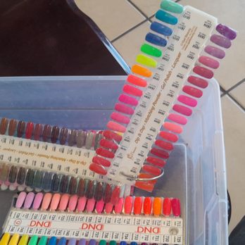 El Paso Nails - Some colors - Multiple plastic strips with various nail polish color samples are organized inside a transparent plastic container. The samples include a wide range of shades from neutrals to bright colors, arranged in rows. The background shows a tiled floor, and part of a person's leg wearing beige clothing is visible on the right side of the image. - Nail Salons Near Me - El Paso, Texas