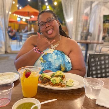 Del Toro - Mango Margarita and chicken and Carne Asada Taco - A woman with glasses and braided hair is sitting at a restaurant table, smiling and sticking out her tongue while making a shaka sign with her hand. The table has a plate of tacos topped with avocado and lime wedges, a yellow cocktail garnished with a strawberry, a small bowl of green sauce, and two cups of water. The background shows a tented outdoor dining area with string lights and other patrons. - Burgers Near Me - Chicago, Illinois