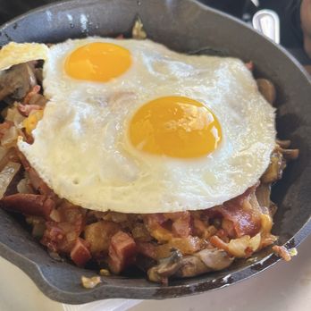Mo's Egg House - Farmhouse Skillet - A black skillet holds two sunny-side-up eggs with runny yolks placed on a bed of cooked bacon, sausage, and vegetables. The dish appears to be part of a breakfast meal, served on a white plate with a napkin underneath. The background suggests a casual dining setting. - Breakfast & Brunch Near Me - Temecula, California
