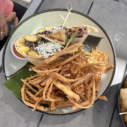 Weekend special breakfast burrito - A plate contains two tacos filled with meat, topped with cheese, and garnished with a leaf, alongside fried plantains on a gray outdoor table.