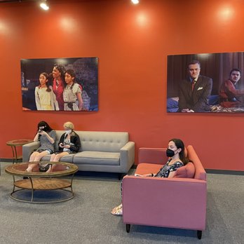 Raven Theatre - Hall - A lobby area of a theater features four people seated on sofas, with two women on the left and one woman on the right, all wearing masks. The seating includes a gray sofa, a pink armchair, and a blue armchair. The wall behind them is painted a reddish color and displays two large framed photographs of theatrical performances, one showing three women and the other showing two men in formal attire. The floor is carpeted, and there are small round tables in front of the sofas. - Summer Camps Near Me - Chicago, Illinois
