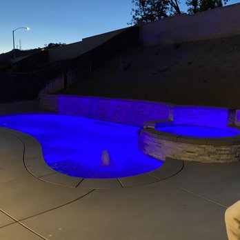 Cerda Pool Construction - The image shows a swimming pool and a hot tub, both illuminated with blue LED lighting, situated outdoors on a concrete deck. The pool has a curved shape with a small fountain feature, and the hot tub is built into a stone surround. The background includes a hillside with a fence and a streetlamp, indicating an outdoor setting during twilight. A person is partially visible in the lower right corner of the image. - Pool & Hot Tub Service Near Me