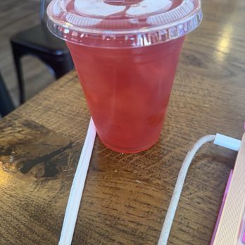 Classic Cups - A pink drink in a clear plastic cup with a sealed lid is placed on a wooden table, with a white straw and a pink laptop nearby. - Coffee & Tea Near Me - Brooklyn, New York