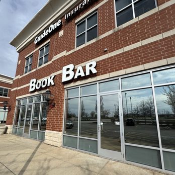 Book Bar - The image shows the exterior of a two-story brick commercial building with large glass storefront windows and doors. The facade displays prominent signage for 'Book Bar' in large white letters and 'GuideOne Insurance' above it. The sidewalk in front is concrete, with shadows cast by nearby objects, and the parking lot reflects in the glass windows, with a few cars and trees visible in the background. - Sandwiches Near Me - Mokena, Illinois
