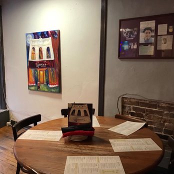 Nonna's - A round wooden dining table is positioned in a restaurant interior, with several menus spread across its surface and a small sign or menu holder in the center. The background includes a white wall with colorful artwork and a framed photo collage, along with a section of exposed brick wall. The setting suggests a casual dining environment with artistic decor. - Italian Near Me - Springfield, Missouri