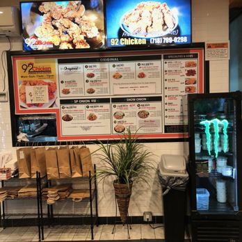 92Chicken - Interior view of 92 Chicken, a Korean-style fried chicken restaurant in Flushing, NY. The image shows a large menu board with various chicken dishes and sides, two digital screens displaying fried chicken images, a rack with takeout paper bags, a potted plant, and a beverage fridge with Monster energy drinks. The setting includes a tiled floor, a trash bin, and visible wiring near the wall. - Korean Near Me - Flushing, New York