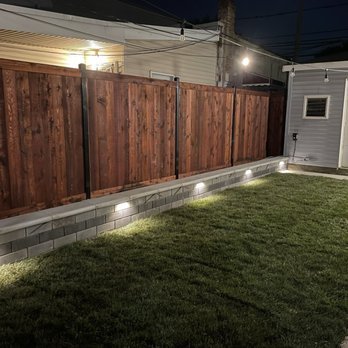Dante's Native Landscape Services - New fencing, retaining wall, sidewalk and sod - A backyard scene at night featuring a wooden privacy fence, landscape lighting along a brick base, and a well-maintained grassy lawn. - Landscaping Near Me