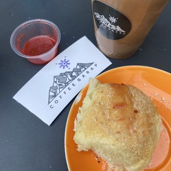 Coffee Sarap - Iced Ube latte and Pandesal with jam - A slice of bread with butter on an orange plate is placed on a table. Next to it, there is a small clear plastic container of jam. A large iced coffee in a plastic cup with a black logo is also on the table. The table surface is dark, and the items are arranged with a napkin featuring the coffee shop's logo. - Coffee & Tea Near Me - Denver, Colorado