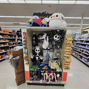 Walgreens - Halloween items - The image shows a store aisle with a display shelf containing Halloween-themed plush toys, decorations, and accessories. The top shelf has large plush toys resembling characters from 'The Nightmare Before Christmas,' including Jack Skellington and Sally, along with a white plush dog with a pumpkin face. The middle section includes smaller plush toys, a decorative wreath, and themed accessories. To the right, there are snack bags, and to the left, a cardboard box with additional items. The background features store shelves with various products, and the ceiling has fluorescent lighting. - Cosmetics & Beauty Supply Near Me - Cedar Park, Texas