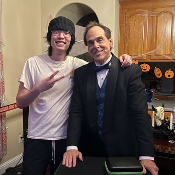 Rappa Magic - Michael Rappa with the birthday boy - A young man with glasses, a beanie, and casual clothing stands next to an older man dressed in a tuxedo with a bow tie, both smiling and posing together. The room features Halloween decorations, including pumpkin cutouts and cobwebs, with a chandelier overhead and wooden cabinets in the background. They are standing behind a table with a black box and a small case on top. - Magicians Near Me - Rocklin, California