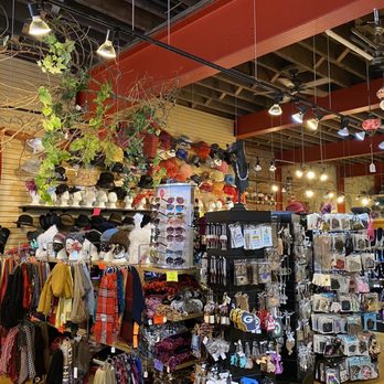 Dimple's - The image shows the interior of a retail store with various accessories including hats, jewelry, scarves, and handbags. Hats are arranged on shelves along the wall, with some hanging from hooks. Jewelry and accessories are displayed on rotating racks and pegboards. The store has overhead track lighting illuminating the merchandise, and the ceiling features exposed beams and ductwork. The background includes a brick wall and slatwall panels for product display. - Women's Clothing Near Me - Racine, Wisconsin
