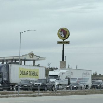 Buc-ee's - The image shows a parking lot with multiple trucks and cars parked in front of a Johnstown sign featuring a lion mascot. The sign is elevated on a pole, with a cloudy sky overhead. In the background, there is a structure with a decorative element and a streetlamp. The scene appears to be taken from a roadside area, with a fence separating the parking lot from the road. - Convenience Stores Near Me - Berthoud, Colorado