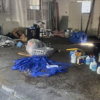 Morpho Carpentry - The image shows a garage with unfinished walls and a concrete floor, cluttered with various items including paint cans, tools, and bags. There are scattered objects and debris on the floor, and a partially open garage door at the top of the image. The space appears used for storage and maintenance activities. - General Contractors Near Me