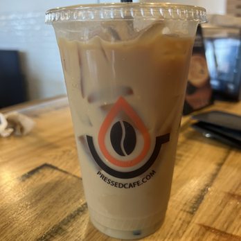 Pressed Cafe - A clear plastic cup containing iced coffee with ice cubes and a black straw, displaying the Pressed Cafe logo with a coffee bean icon and website. The cup is on a wooden table inside a cafe, with a blurred background showing part of the cafe interior and some objects on the table. - Juice Bars & Smoothies Near Me - Newton, Massachusetts