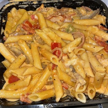 Scratch Kitchen - Pasta Primavera Dinner - A black plastic takeout container contains pasta primavera with cooked penne pasta, cherry tomatoes, sliced onions, green peppers, and a creamy sauce. The dish appears freshly prepared, with visible vegetables and sauce coating the pasta. The container is open, showing the food inside, and some sauce has spilled onto the edges. - Breakfast & Brunch Near Me - Olney, Maryland