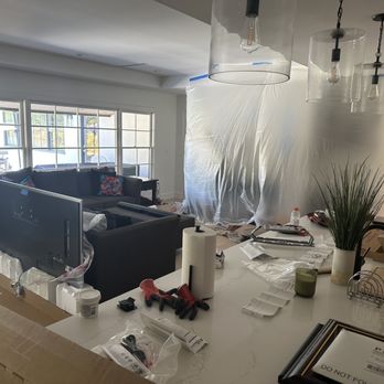 Marvel Audio - Paule is a genius and made sure not to create a mess by setting up a covered work area. - The image shows a living room with large windows allowing natural light, a black sectional sofa with colorful pillows, and a plastic sheet covering part of the wall, suggesting renovation. The foreground features a kitchen counter cluttered with tools, paper towels, a plant, bananas, and various construction materials, indicating ongoing remodeling activities. - Home Theatre Installation Near Me - Folsom, California