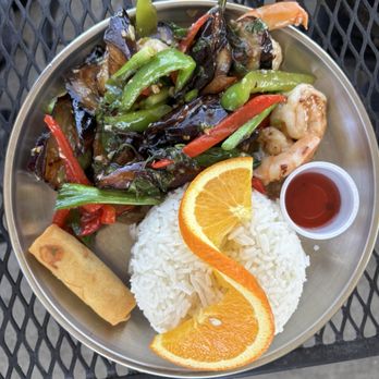 Thai Hut - Basil eggplant added shrimp. - The image shows a stainless steel plate with a serving of white rice topped with orange slices. Next to the rice, there is a portion of stir-fried vegetables including bell peppers, eggplant, and green beans, with shrimp mixed in. A spring roll is placed on the side, and a small cup of red sauce is also present. The plate is on a black metal outdoor table. - Thai Near Me - Redding, California