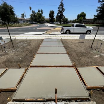 Jimenez Concrete - Front walk - Concrete sidewalk sections are in the process of being poured and set on a construction site in front of a building. The site shows freshly poured concrete slabs with wooden forms, and the surrounding area includes dirt and construction materials. A street with a parked white pickup truck and trees is visible beyond the construction zone, under a clear blue sky. - Masonry/Concrete Near Me