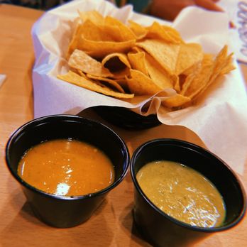 Sagrado - Chips and Salsa - A basket of tortilla chips lined with white paper is placed on a wooden table, accompanied by two black cups of salsa, one orange and one green. The background shows a person holding a menu and part of a restaurant setting. The image captures a typical Mexican appetizer presentation. - Mexican Near Me - Waco, Texas