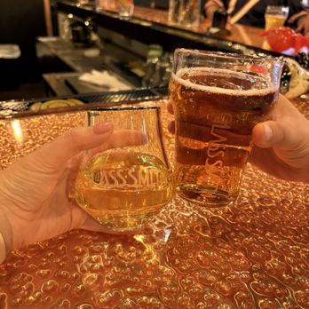 The Glass Smith - Custom glassware - Two glasses of beer are being held up at a bar with a copper-colored, textured countertop. The background shows a bartender and bar equipment. - Bars Near Me - Richmond, Illinois