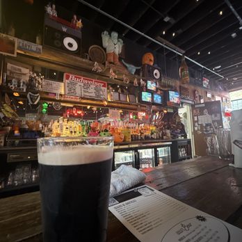 Broken Hearts - A pint of dark beer with a foamy head is placed on a wooden bar counter inside a bar with various decorations and a busy background. - Wine Bars Near Me - Chicago, Illinois