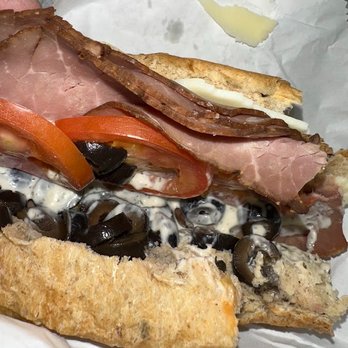 Subway - "Just tomatoes & Ranch dressing." (got olives with my roast beef - not a very yummy combo) - A close-up of a sandwich with layers of sliced ham, roast beef, tomato, and black olives, topped with mayonnaise on a crusty baguette-style bread. The sandwich is partially wrapped in white paper, with some butter visible on the bread. - Sandwiches Near Me - Madison, Wisconsin