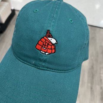 Target - The image shows a teal-colored baseball cap with an embroidered depiction of Snoopy, the cartoon dog, dressed in a red jacket and a red hat. The cap is held in a person's hand, with a blurred background that appears to be inside a retail store or similar setting. The cap has visible stitching and ventilation eyelets on the crown. - Department Stores Near Me - Skokie, Illinois