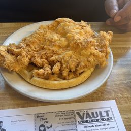 Chicken & Waffle - Fried chicken served on a waffle is placed on a white plate on a wooden table, with a person's hand visible in the background.