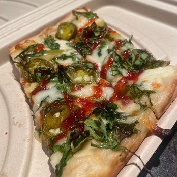 Bartlet Street - Isaac Newton (partially consumed) FABULOUS!! - A rectangular slice of pizza features toppings including sliced jalapeños, leafy greens, melted cheese, and red sauce. The pizza is placed in a beige takeout container, with a dark surface underneath. The toppings are spread evenly, and the cheese appears melted and slightly browned. - Pizza Near Me - Andover, Massachusetts