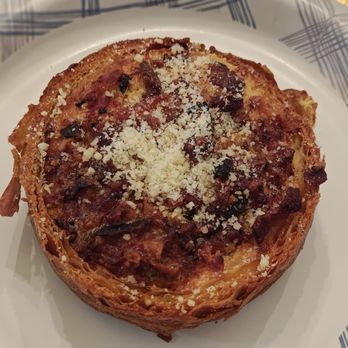 Baker 305 - Bacon egg and cheese quiche - A baked pastry with a golden-brown, flaky crust, topped with melted cheese, small pieces of bacon or ham, and herbs. The pastry is presented on a white plate with a blue pattern, and the image captures the detailed texture of the toppings and crust. - Bakeries Near Me - Miami, Florida