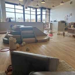 The image shows an indoor children's play space with a small slide, soft toys, and scattered play equipment on a wooden floor. Large windows in the background let in natural light, and the ceiling has multiple ceiling lights. The space includes a reception or activity area with desks and wall decorations, indicating a play or learning environment for children.