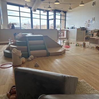Giggles Play - The image shows an indoor children's play space with a small slide, soft toys, and scattered play equipment on a wooden floor. Large windows in the background let in natural light, and the ceiling has multiple ceiling lights. The space includes a reception or activity area with desks and wall decorations, indicating a play or learning environment for children. - Indoor Playcenter Near Me - Highwood, Illinois