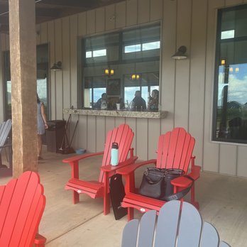 Deep Sky Vineyard - Outdoor window where tastings can be poured - Outdoor patio area featuring red and gray Adirondack chairs, with a small table holding a water bottle and a bag. The building has a large window with a granite ledge, through which people are visible inside, possibly at a bar or counter. The patio has a wooden ceiling and vertical siding, with a trash can and cleaning tools nearby. - Wine Tasting Room Near Me - Elgin, Arizona