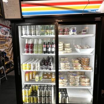 The Nitro Bar- Providence - Grab and go items - A black refrigerated display case with a glass door showcases various bottled drinks, including sodas, juices, and energy drinks, along with pre-packaged salads and desserts on multiple shelves. The case is located indoors, with a menu or sign attached to the top left corner and some plants visible above it. The setting appears to be a convenience store, café, or similar retail environment. - Coffee & Tea Near Me - Providence, Rhode Island