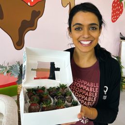 Adult Class - 12 strawberries - A woman with dark hair and a nose piercing holds a white box filled with chocolate-covered strawberries, smiling at the camera.
