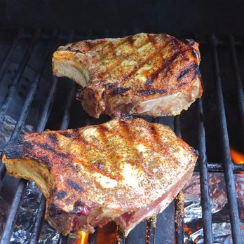 Raley's - grilled up great - Two pork chops are cooking on a barbecue grill, showing grill marks and a seasoned surface, with flames and foil underneath. - Grocery Near Me - Redding, California