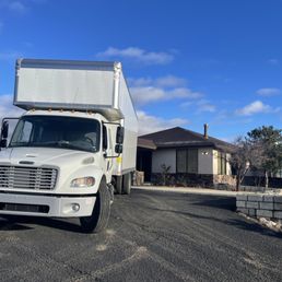 2 truck move- Prescott to Phoenix - A white moving truck is positioned on a paved driveway in front of a single-story house with a dark roof and stone exterior accents. The truck's cab is visible, and the sky above is bright blue with some scattered clouds. The surrounding landscape includes a few trees and a low stone wall.