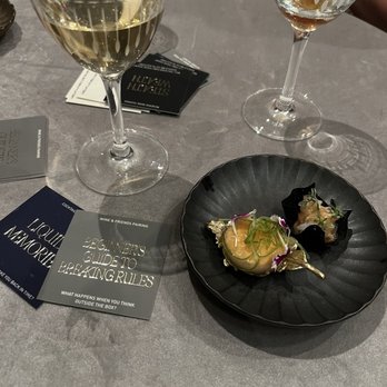 Class Act - Two glasses of white wine and a black plate with two small appetizers are on a gray table, with business cards and cards nearby. - New American Near Me - Chicago, Illinois